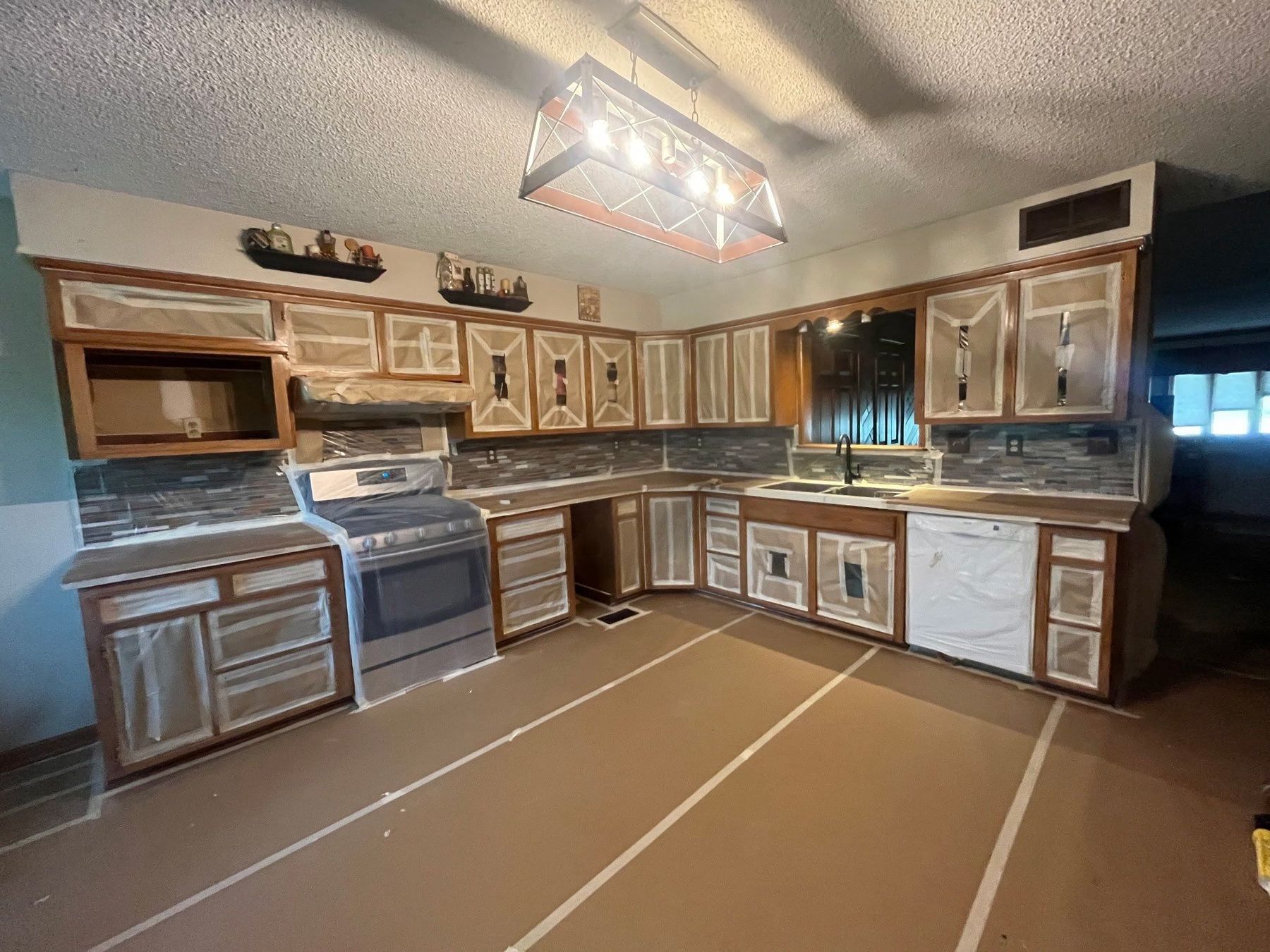 Kitchen prepped for cabinet painting — floors papered, doors removed, boxes and appliances fully masked